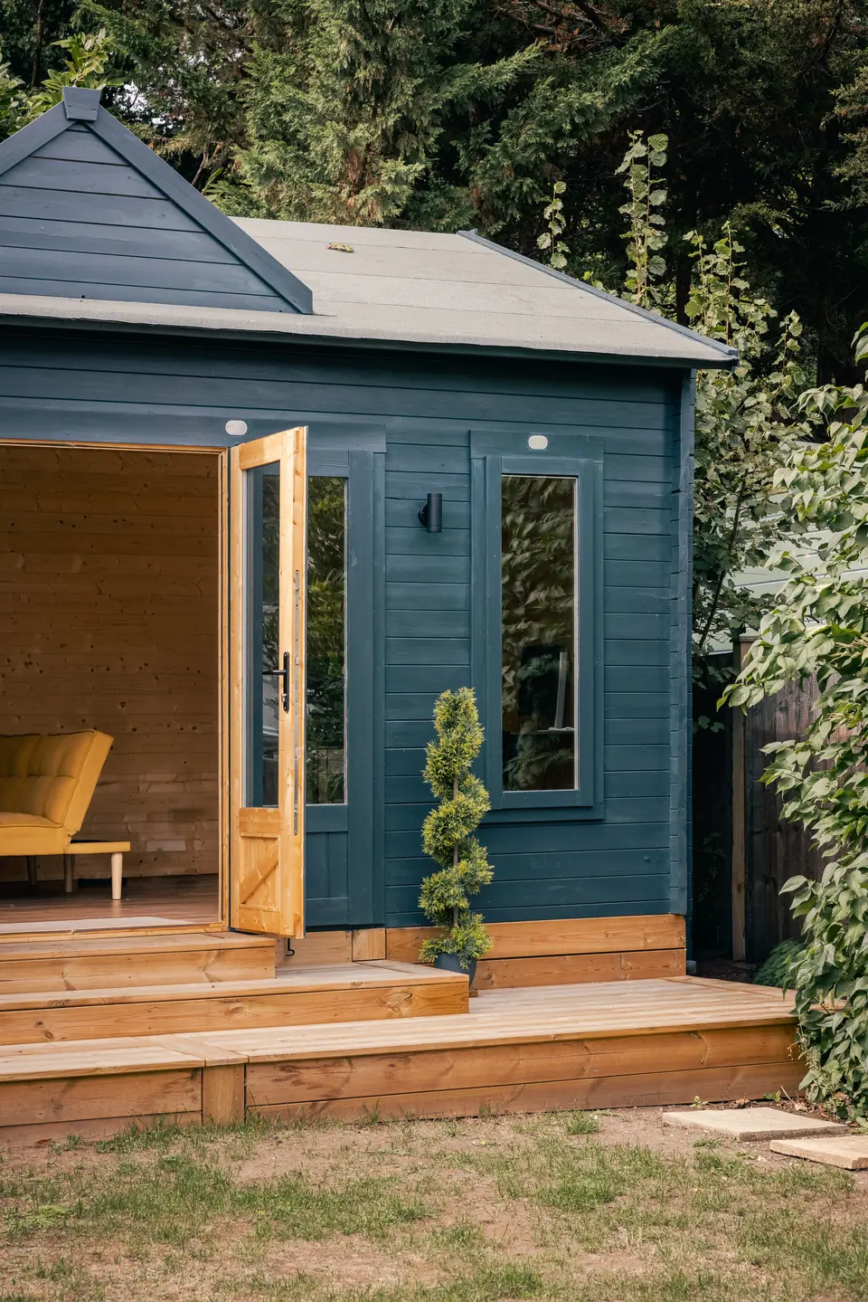 Side view of the navy garden room, its deck and open door beside a tall hedge