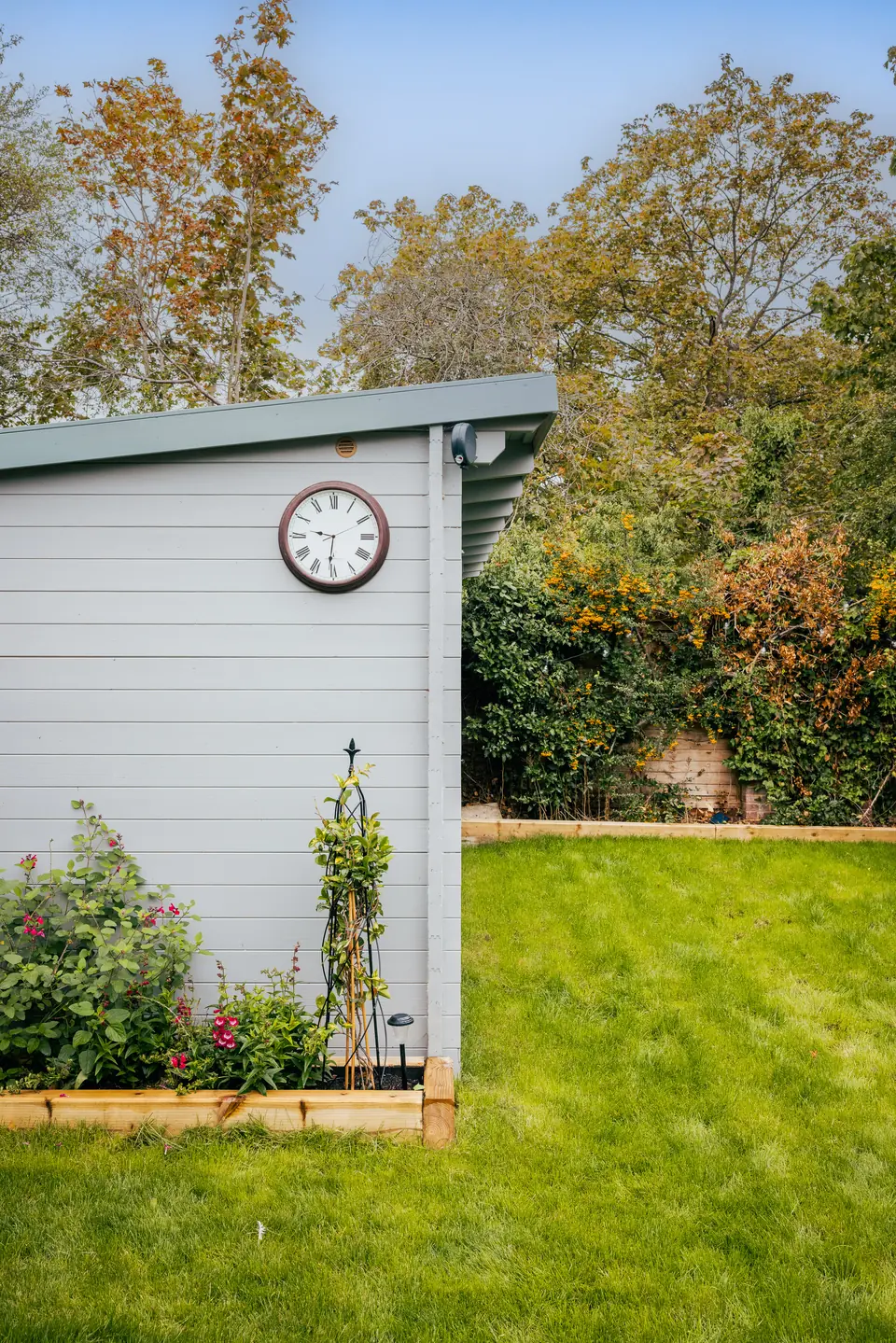 Gable end of the sage log cabin with a clock, climbing roses and a timber raised bed
